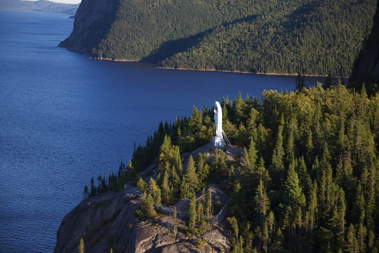 Statue blanche sur un promontoire rocheux surplombant l'eau dans la région du Saguenay-Lac-Saint-Jean.