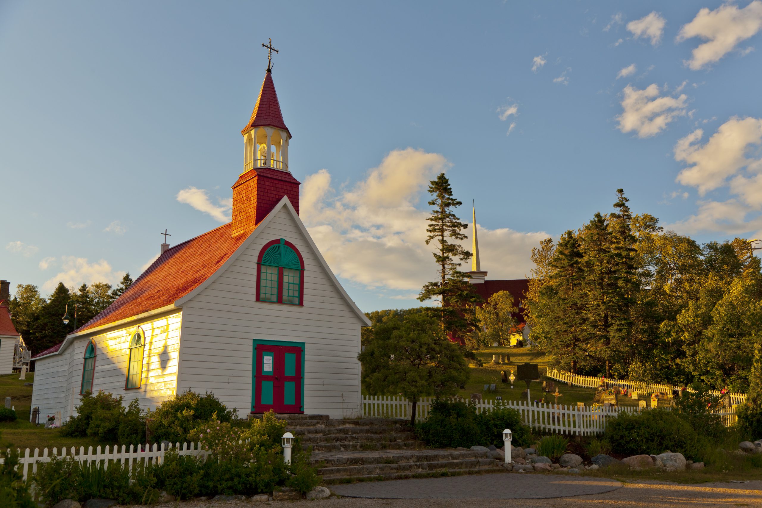 Petite église blanche avec toit rouge et porte turquoise en Côte-Nord, Québec, sous un ciel nuageux en fin de journée.
