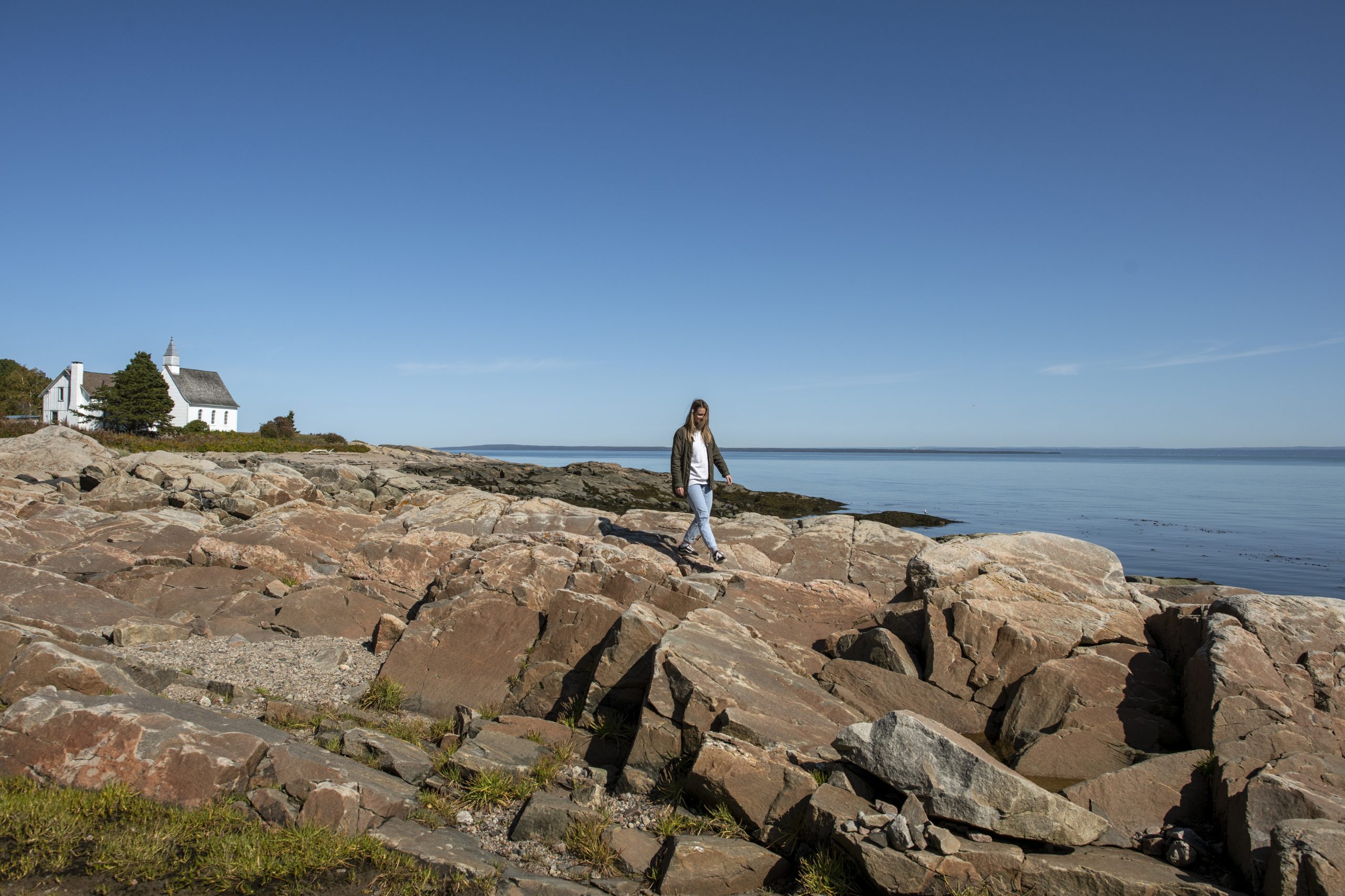 Femme marchant sur un rivage rocheux avec une petite église blanche en arrière-plan, près de la mer sous un ciel bleu.